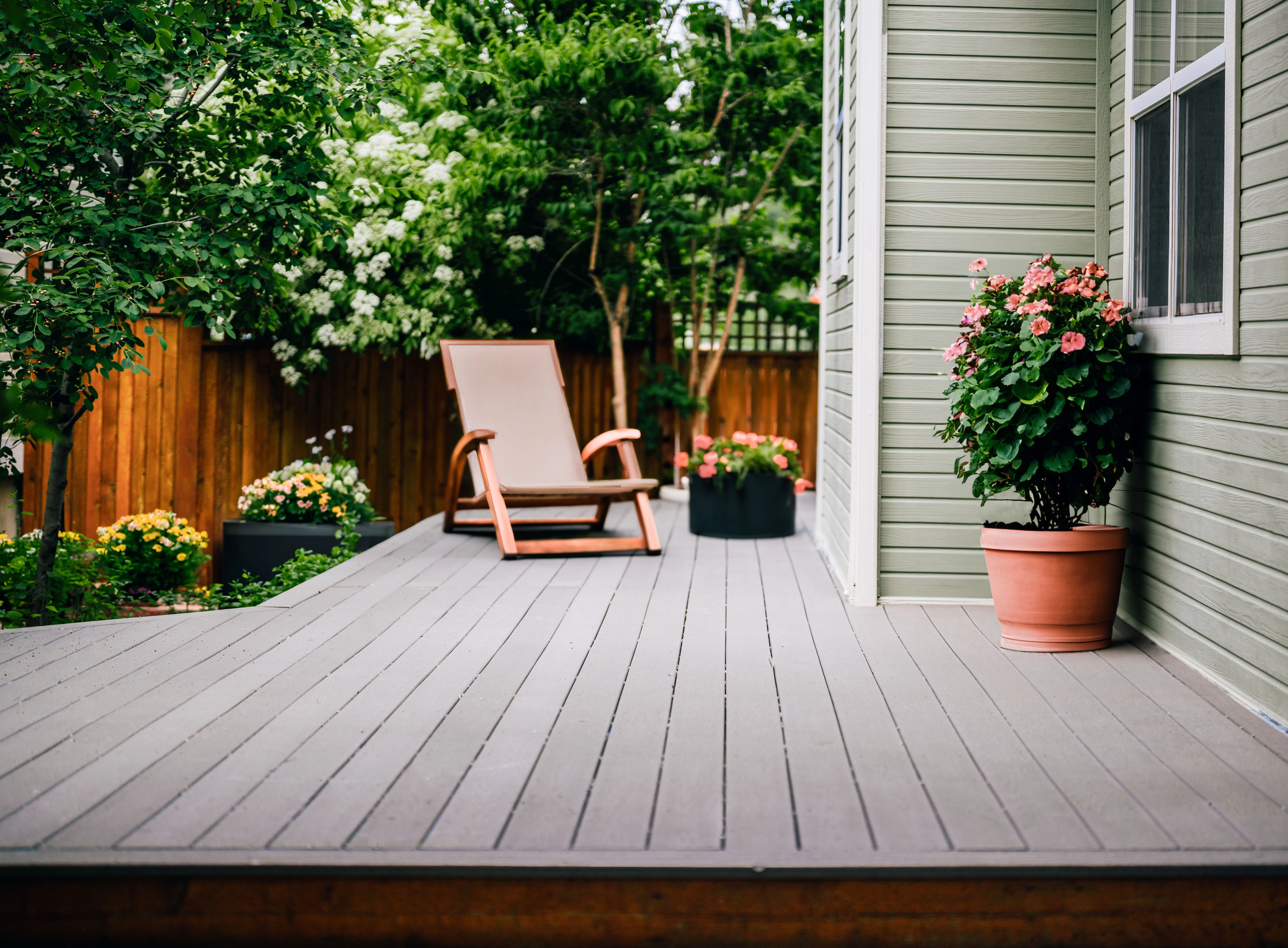 Gray painted deck without a railing surrounded by flowers and trees. House siding is light green. 