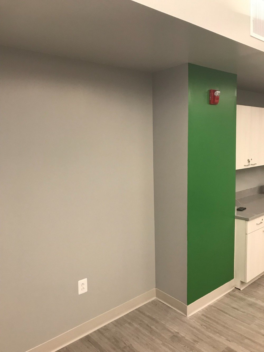 Gray wall alongside a bright green wall next to a kitchenette with gray wood looking flooring in a commercial workspace