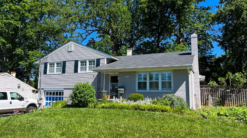 This image shows the exterior of a light gray brick house with a chimney on the right side. The house has a gable roof with shingles and features large windows with white trim on the left side.