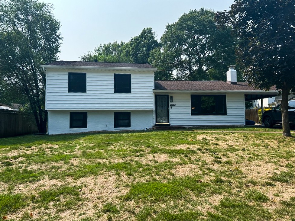 The house in the photo showcases a newly painted exterior with bright white siding and black window frames, giving it a modern and sleek look. The fresh paint enhances the home's clean and updated appearance against a backdrop of lush trees.