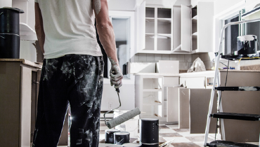 Photo of a man covered in paint and holding a used paint roller in a freshly painted white kitchen.