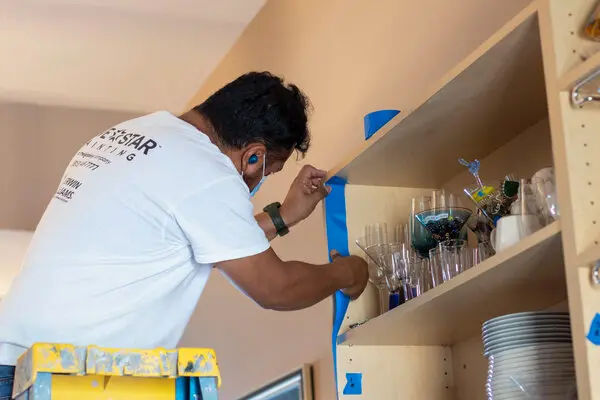 Male Five Star Painting technician applying blue painter's tape to bookshelf with wineglasses on shelf.