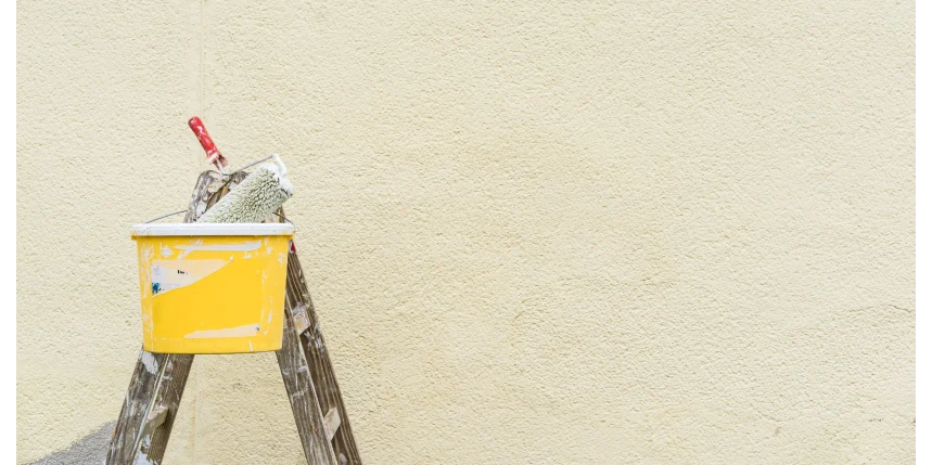 A paint roller sits in a bucket on top of a ladder in front on a painted exterior wall.