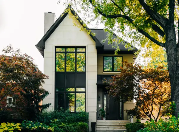 Exterior of modern home with beige panel siding, black roof, trim, and door, and large windows. Home is surrounded by trees with shrubbery near the entrance.